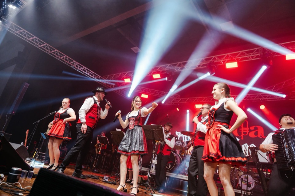 Mitglieder der Banda Original in Tracht singen und tanzen auf der Bühne im Scheinwerferlicht.