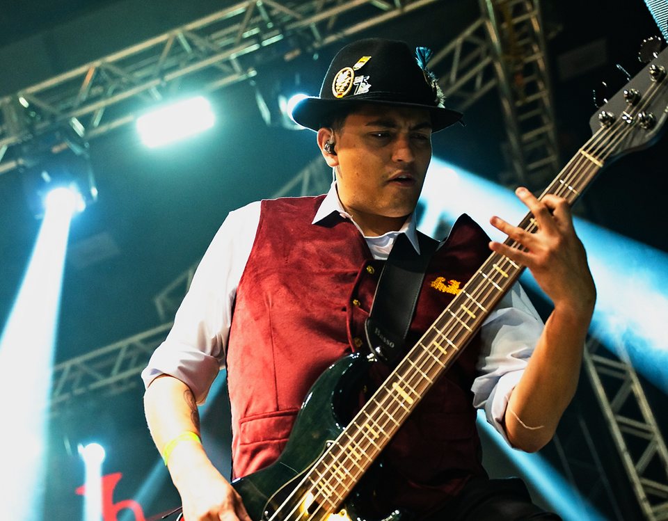 Bassist mit Hut und weinroter Weste spielt auf der Bühne unter Lichtstrahlen.
